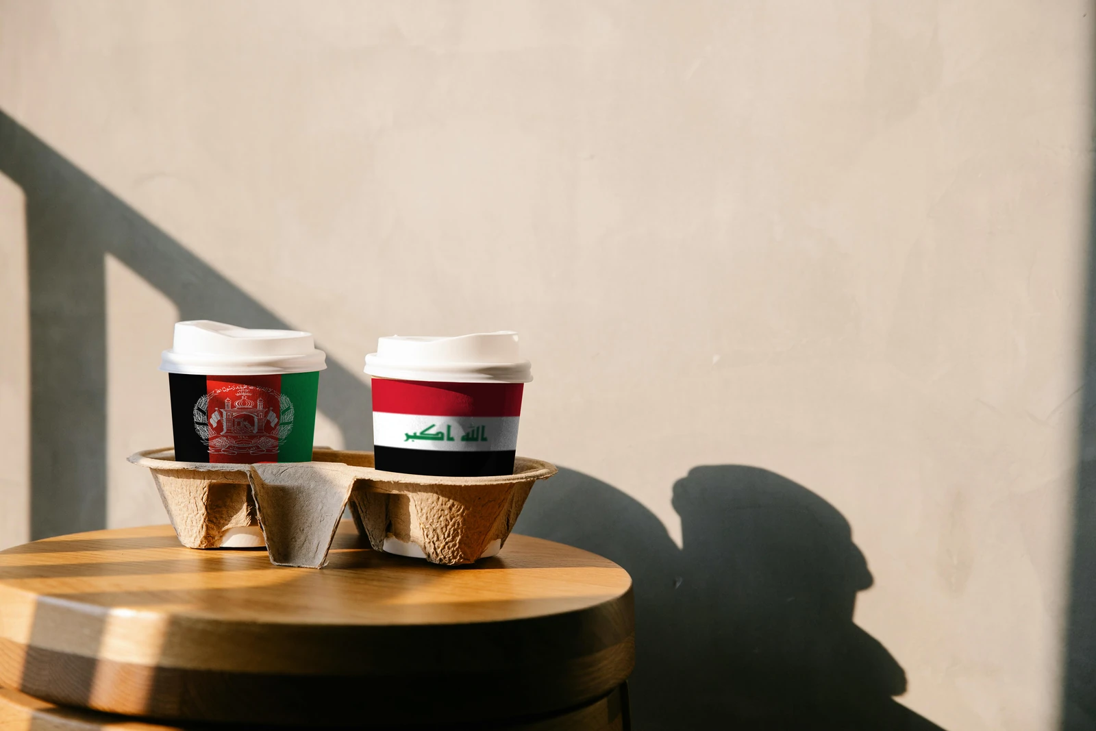 Two takeaway cups in a cardboard carrier on a round wooden table: one printed with the flag of Afghanistan, the other with the flag of Iraq