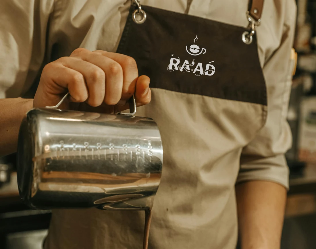 A barista in a brown RA’AD apron holding a steel milk pitcher