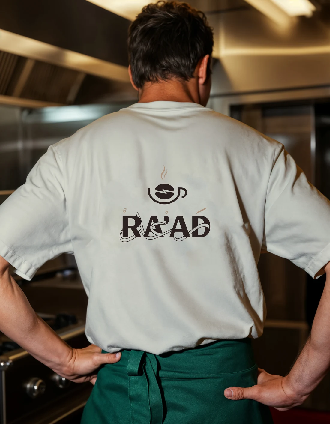 A RA’AD team member from behind in a white T-shirt with the RA’AD logo and a green apron, in a steel kitchen